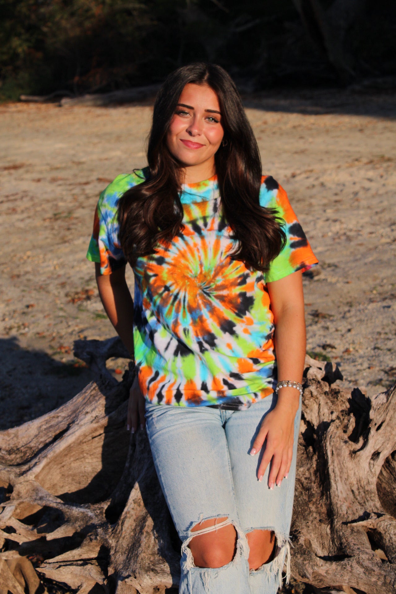 Person wearing a tie-dye shirt sitting on a rock by a body of water.