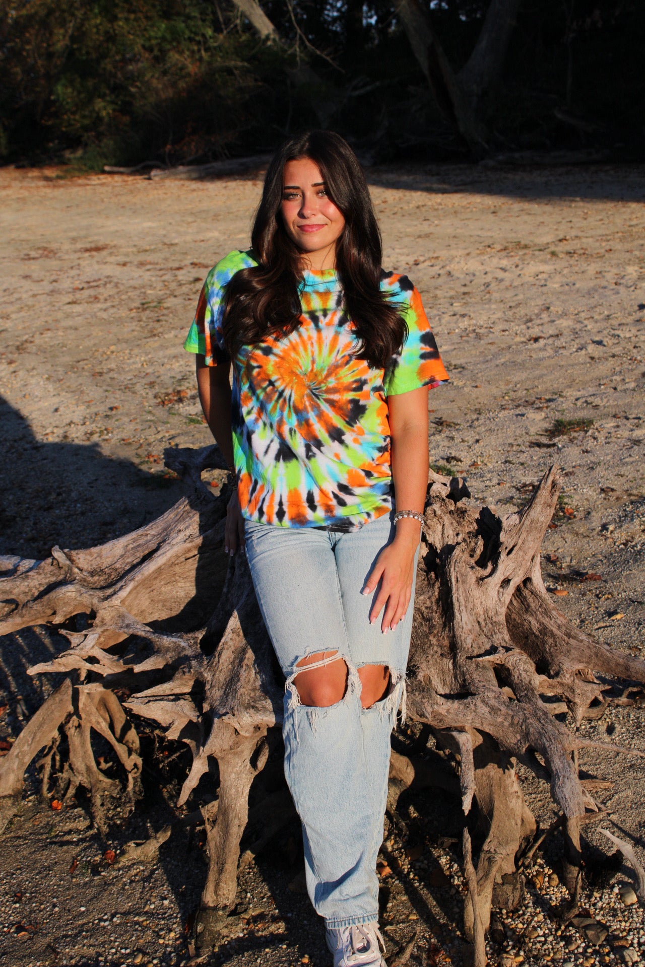 Person wearing a tie-dye shirt and jeans standing on a beach with driftwood.
